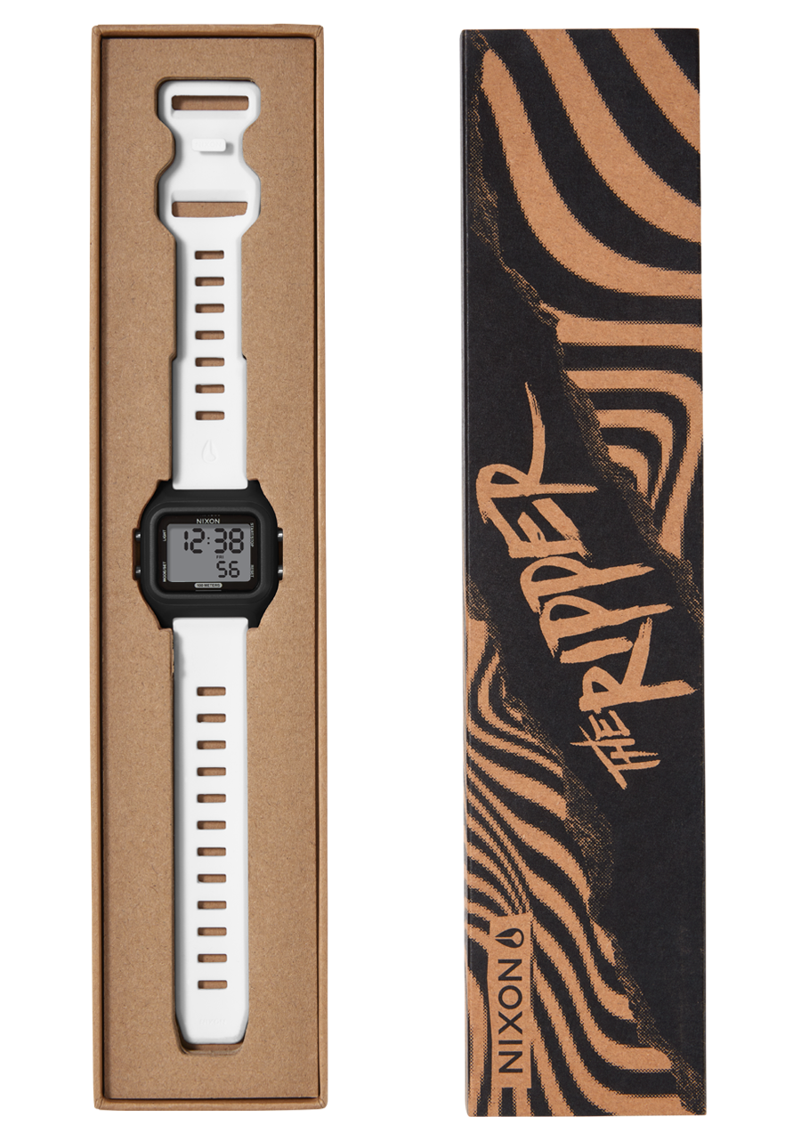A white Nixon digital watch with a black face and white strap is presented in an open brown cardboard box next to its black and brown striped packaging that reads "THE RIPPER" and "NIXON".