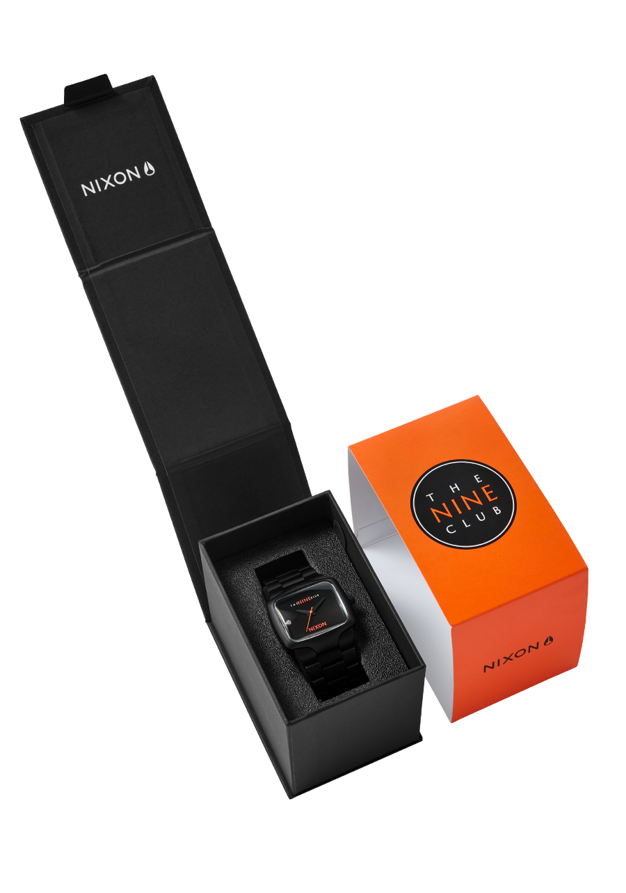 A black Nixon watch with an orange accent on the face is presented in an open black box, next to an orange box with "The Nine Club" logo.