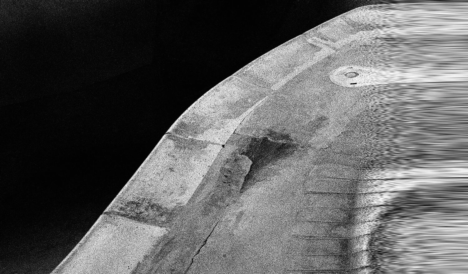 An abstract, black and white image features a curved, textured concrete-like surface with a small circular detail, set against a dark background.